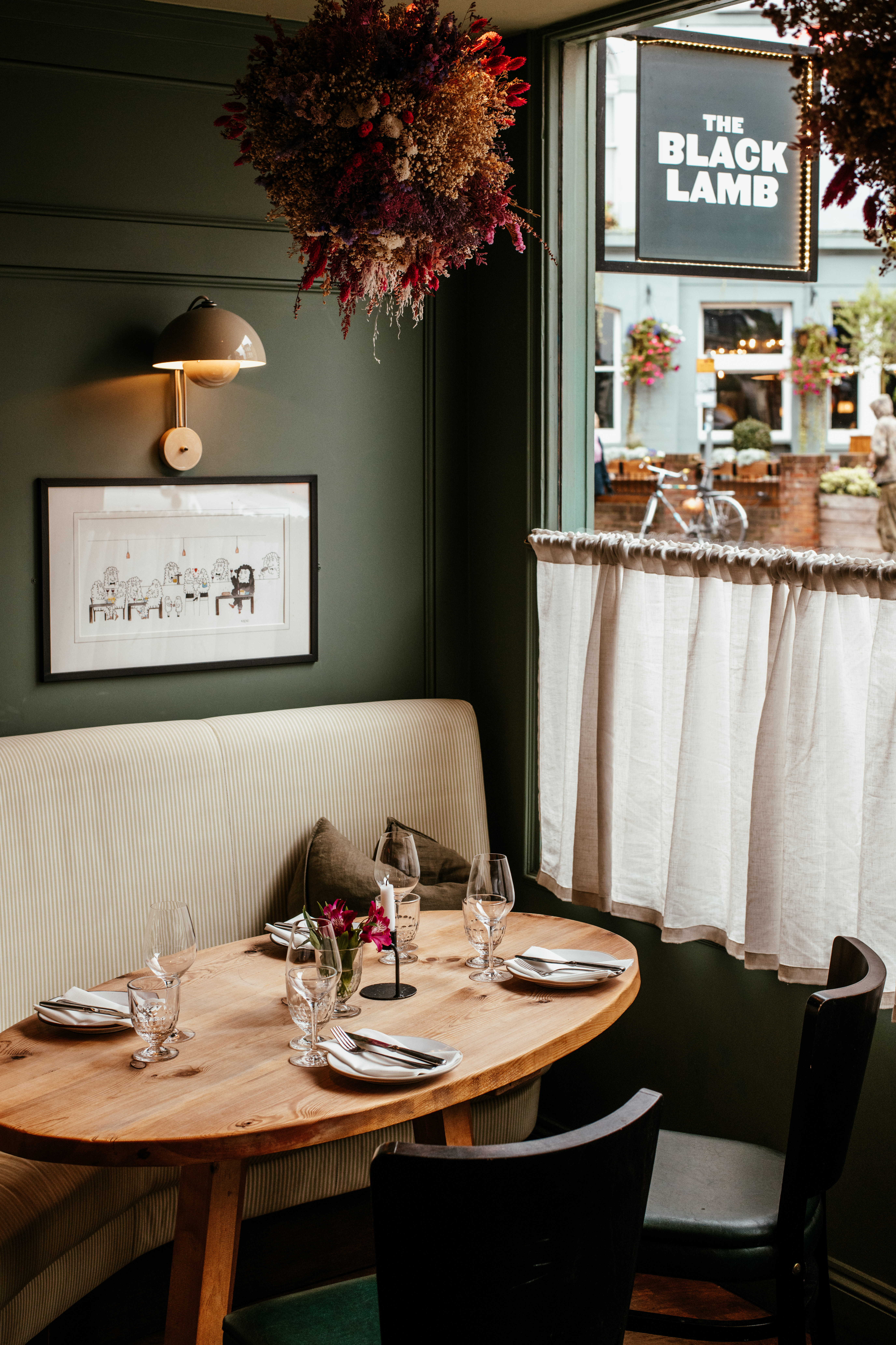 A corner table inside The Black Lamb in Wimbledon Village, with green walls, soft lighting and the restaurant sign visible through the window.